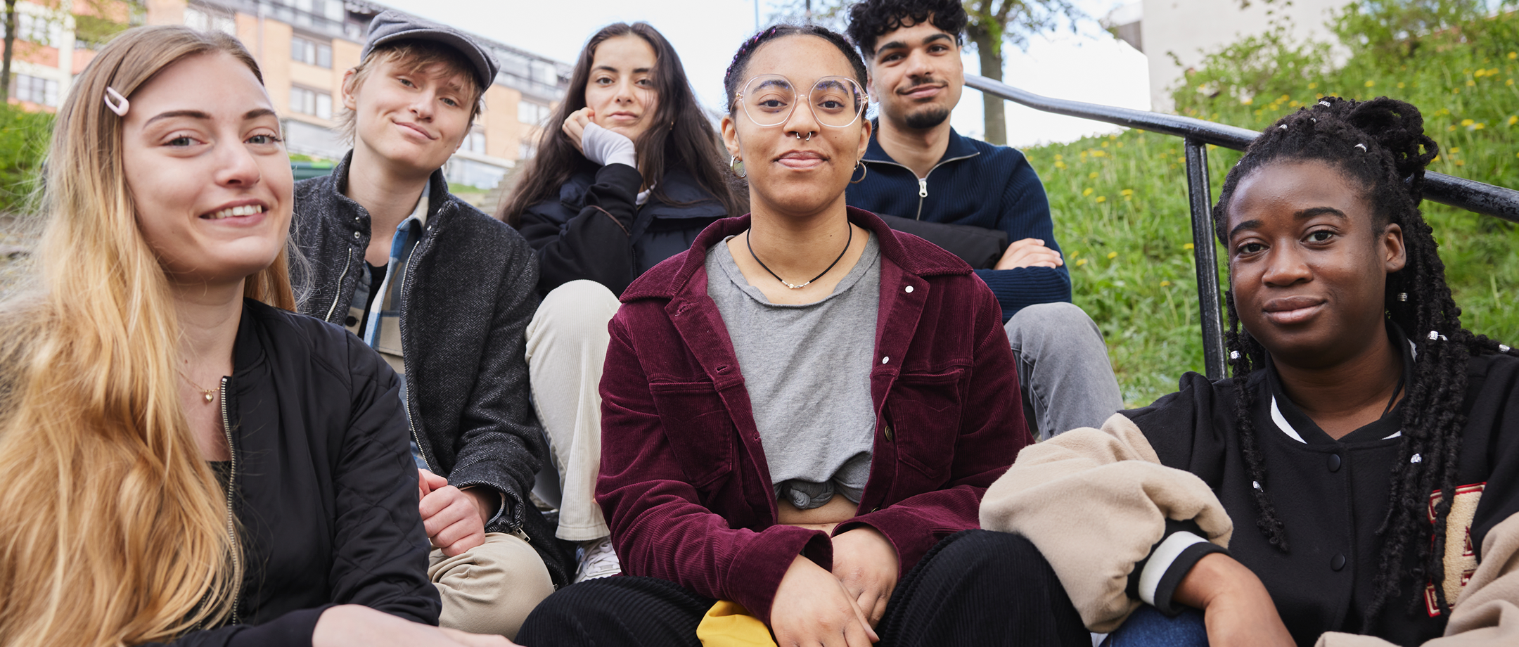 En grupp ungdomar sitter utomhus och tittar in i kameran
