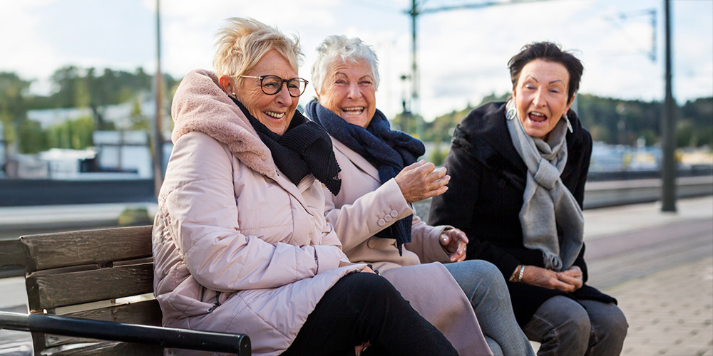 Fyra äldre damer sitter på en bänk på en tågperrong och skrattar.