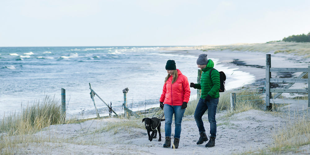 En man och en kvinna promenerar längs med en strand tillsammans med en mellanstor hund.